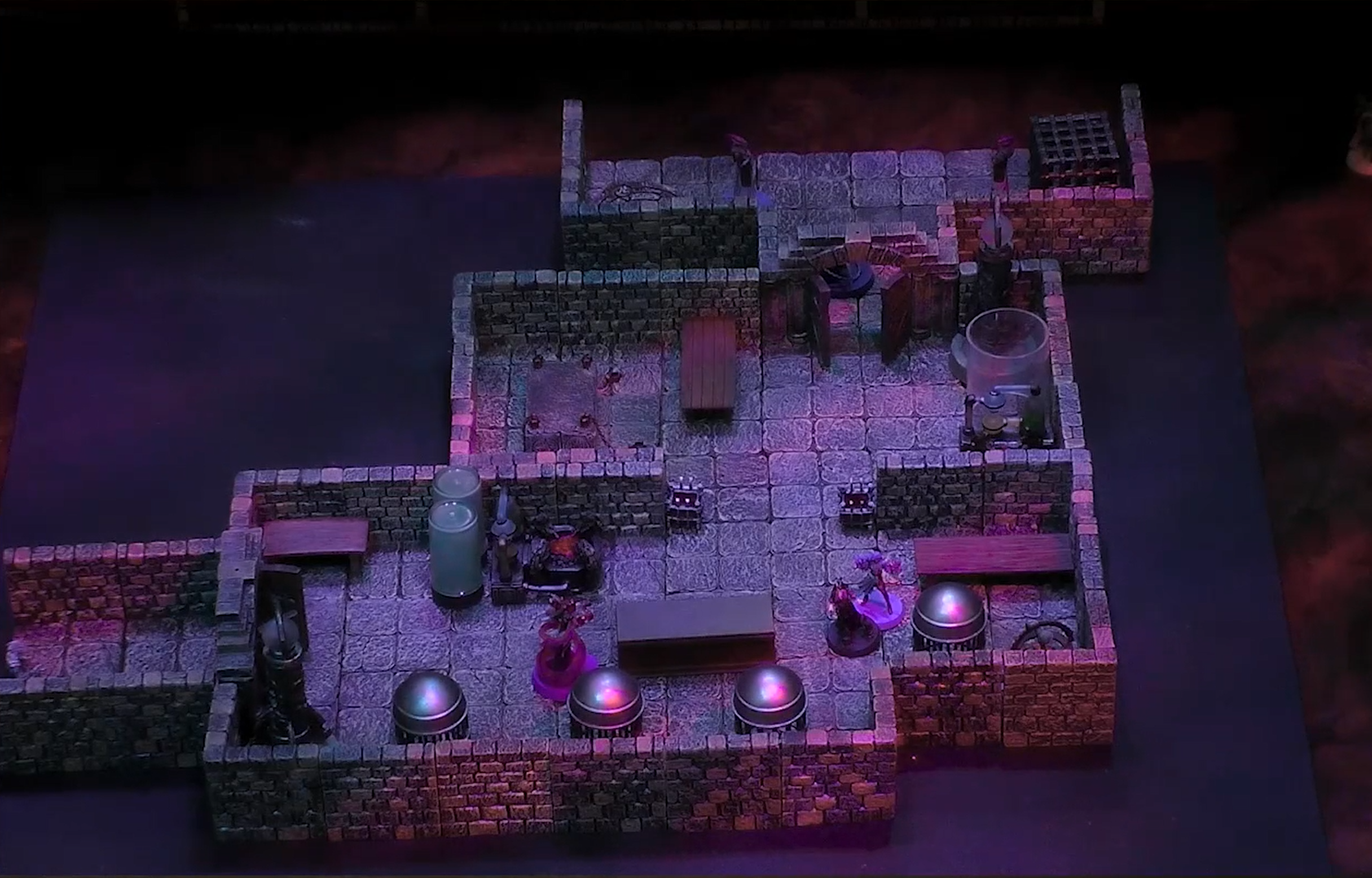 A battlemap of a multi-roomed interior space containing various large cylindrical glass tubes. Two Reilorans are through a door near the top by a large iron cage. Imogen and Laudna are across a table from a Reiloran Mystic surrounded by a purple swirl.