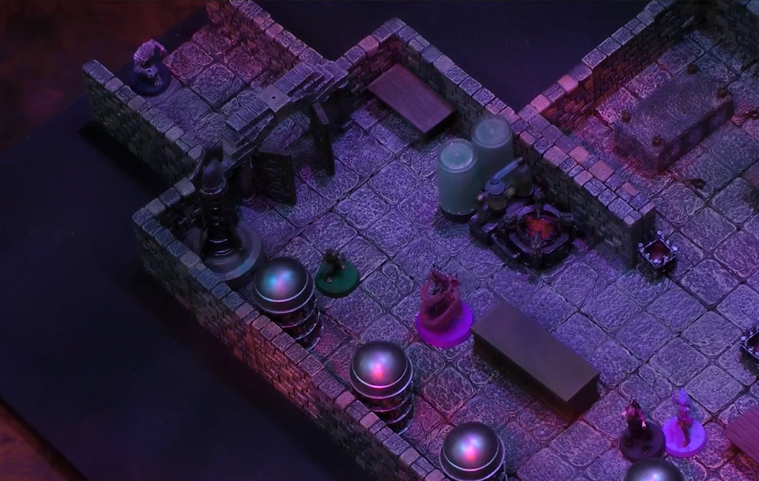 A battlemap of a stone room containing two large cylindrical glass tubes, a forge-like apparatus, and a line of round metal cages against the wall. Imogen and Laudna are across a table from a Reiloran Mystic surrounded by a purple swirl. Orym is behind them, and further back through an open door is Chetney in wolf form.