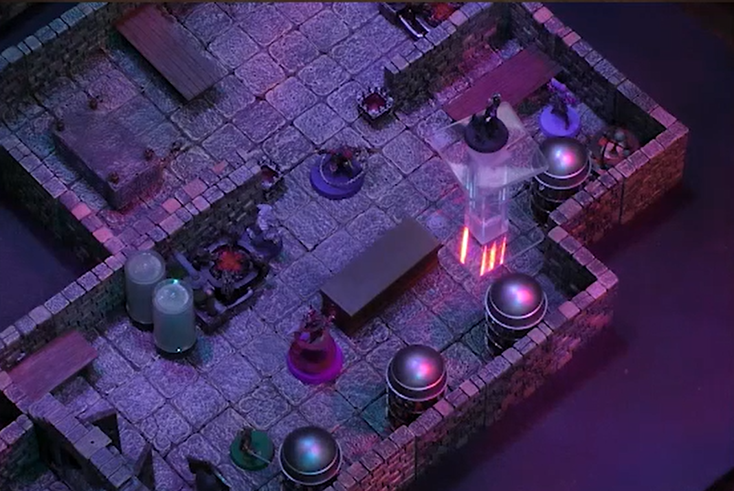 A battlemap of a stone room containing two large cylindrical glass tubes, a forge-like apparatus, and a line of round metal cages against the wall. Imogen is by the wall near a table and Laudna is on a transparent token platform not far in front of her across a table from a Reiloran Mystic surrounded by a purple swirl. Orym is behind them and a Reiloran Shrike is in the entrance to the room.