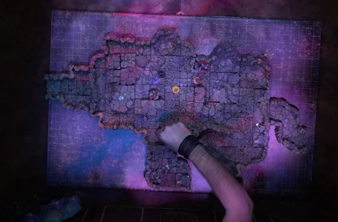 An overhead shot of a battlemap of a large cave chamber scattered with collections of cloudy white crystals. Ashton, Imogen, Laudna, Fearne, and FCG are standing in frame. Matt’s arm reaches in to move a token.