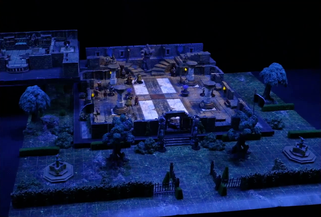 A battlemap of a stone palace split into two levels, surrounded by a courtyard. Downstairs is a marble floor, marble pillars, and twin staircases curving up on either side of a large robed statue holding a lantern and open book. A number of people are scattered throughout the downstairs space. Upstairs is a hallway leading past a room with a bearskin rug, wooden table, and balcony. A number of robed individuals including Ethrand Tachonis gather outside the room, down the hall from a fallen guard. Two black-clothed individuals stand outside the entrance to the palace next to two pillars topped with large spherical crystals. The rest of the courtyard contains trees, two fountains, and an open gate leading in.