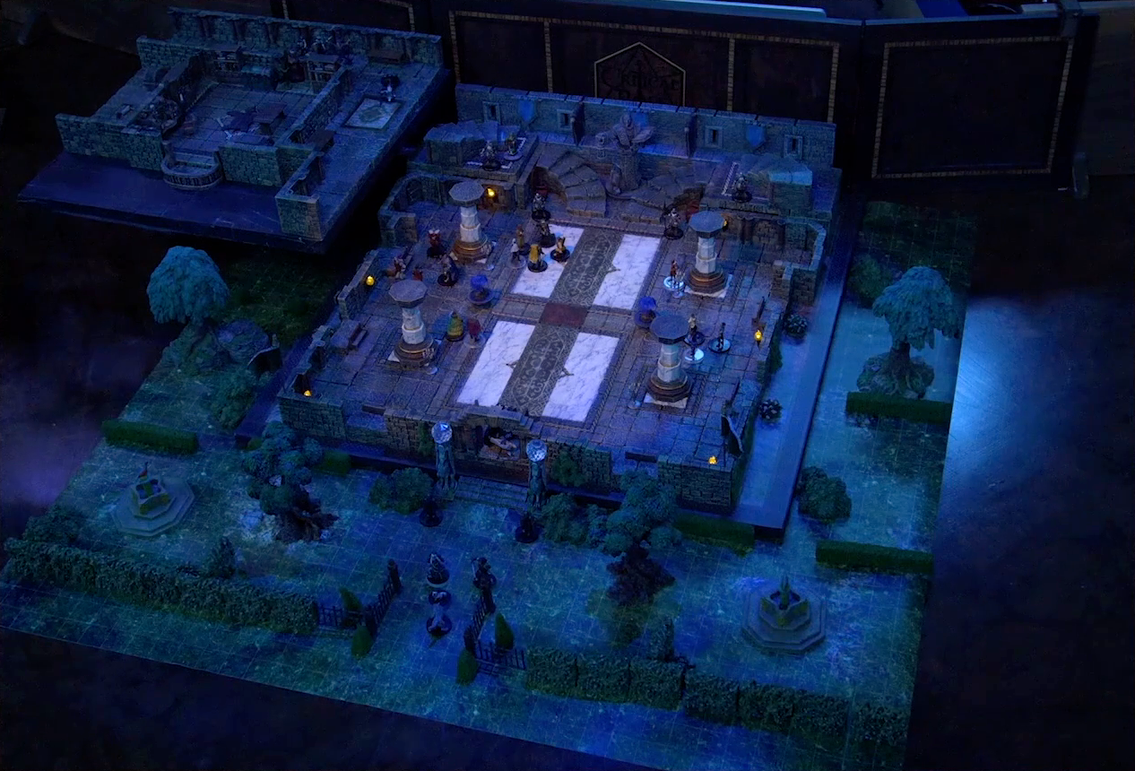 A battlemap of a stone palace split into two levels, surrounded by a courtyard. Vaelus, Julien, Pincushion, and Thaisha in wolf form stand outside the palace entrance, just inside the open gate. Downstairs is a marble floor, marble pillars, and twin staircases curving up on either side of a large robed statue holding a lantern and open book. A number of people are scattered throughout the downstairs space. Upstairs is a hallway leading past a room with a bearskin rug, wooden table, and balcony. A number of robed individuals including Ethrand Tachonis gather outside the room, down the hall from a fallen guard. Two black-clothed individuals stand outside the entrance to the palace next to two pillars topped with large spherical crystals. The rest of the courtyard contains trees, two fountains, and an open gate leading in.