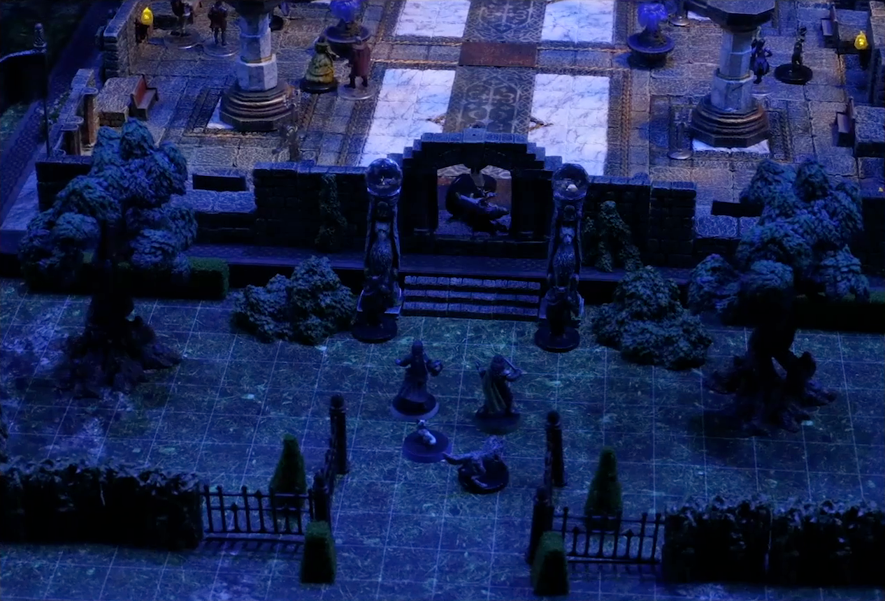 A battlemap of a stone palace surrounded by a courtyard. Vaelus, Julien, Pincushion, and Thaisha in wolf form stand outside the palace entrance, just inside the open gate. Downstairs is a marble floor, marble pillars, and number of scattered people. Two black-clothed individuals stand outside the entrance to the palace next to two pillars topped with large spherical crystals. Primus Tachonis stands just inside the entrance over a fallen guard.