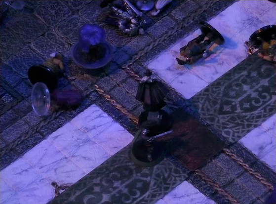 A battlemap of a stone palace focused on Julien facing off against Primus Tachonis. Primus wears long black robes and stands facing away from Julien, who holds out his sword. A number of individuals are laying on the ground nearby on the marble floor. A large partially-melted black candle stands in front of Primus.