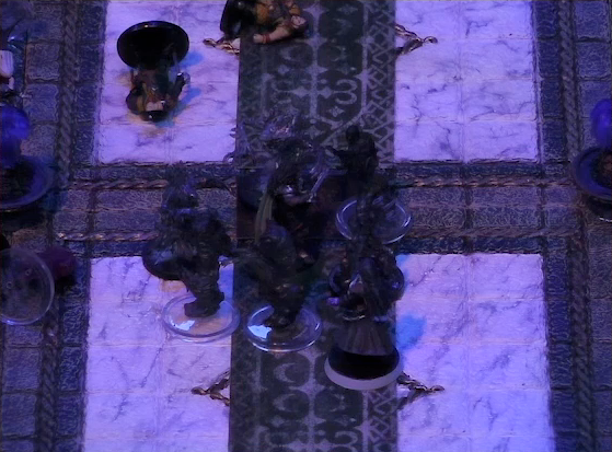 A battlemap of a stone palace focused on Julien surrounded by a number of robed individuals and humanoid creatures, with Vaelus fighting from outside the mob. A number of individuals are laying on the ground nearby on the marble floor. Two large partially-melted black candles stand a short distance from the center of the floor.