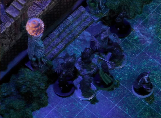 A battlemap of a stone palace courtyard focused on the palace entrance. Julien and Vaelus face off against five tree-like monsters and two black-clothed individuals. Two stone pillars bearing carvings of griffins and topped with large spherical crystals frame the entryway. One crystal glows with an eerie orange light.