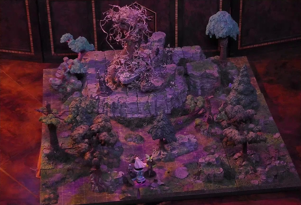 A battlemap of the Wraith Tree on a large rocky mound in a sparse forested space. Kattigan, Wicander, Teor, Thimble, Wulferic, and Tyranny look towards the mound from the edge of the battlemap. The Wraith Tree has thin, white, spaghetti-like tendrils hanging from its bare branches. Three small wooden structures covered in skulls surround the tree.