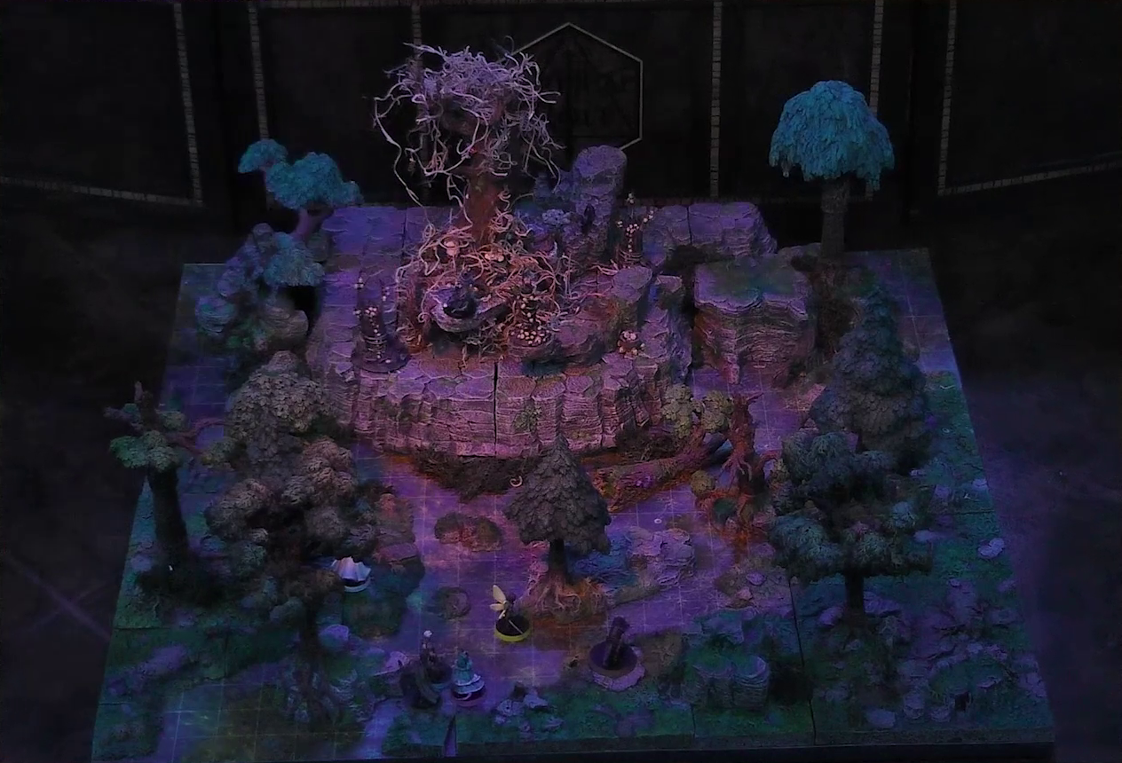 A battlemap of the Wraith Tree on a large rocky mound in a sparse forested space. Kattigan, Wicander, Teor, Thimble, Wulferic, and Tyranny look towards the mound from the edge of the battlemap. The Wraith Tree has thin, white, spaghetti-like tendrils hanging from its bare branches. A black-cloaked specter stands at the base of the tree. Three small wooden structures covered in skulls surround the tree.