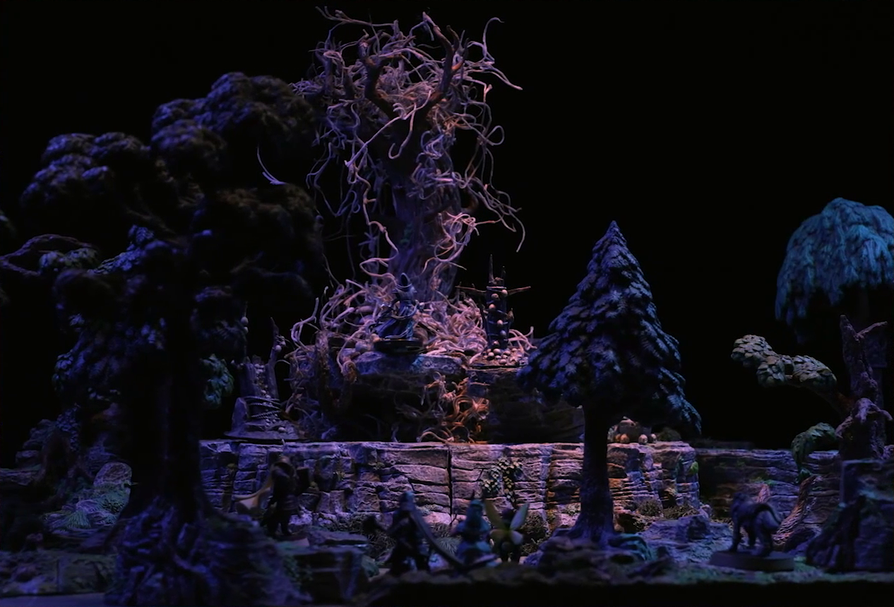 A battlemap of the Wraith Tree on a large rocky mound in a sparse forested space. The Wraith Tree has thin, white, spaghetti-like tendrils hanging from its bare branches. A black-cloaked specter stands at the base of the tree. Wooden structures covered in skulls surround the tree. Kattigan, Wicander, Teor, Thimble, Wulferic, and Tyranny look towards the mound, blurry in the foreground.
