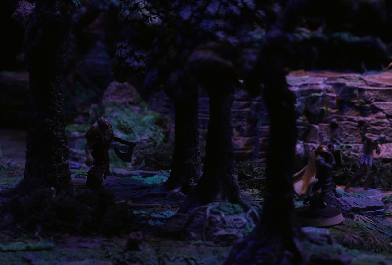 A battlemap of a sparse forested space focused on a zombie wielding an axe standing in the shadow of some trees. It approaches Teor from behind as he faces off against another, thinner zombie. The base of a large rocky mound is visible in the background.