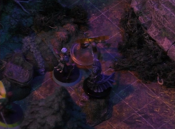 A battlemap of a sparse forested space focused on Wicander facing off against a zombie and a black-cloaked specter. An orange condition ring is around the zombie. Thimble flies a short distance away at the edge of the image. The mossy base of a large rocky mound is visible in the background.