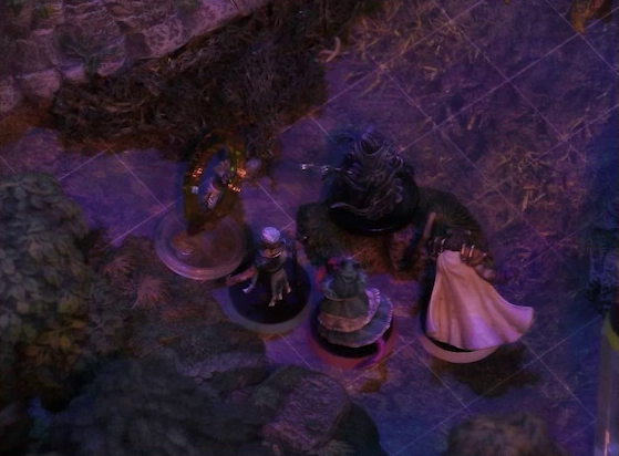 A battlemap of a sparse forested space focused on Wicander, Teor, and Tyranny facing off against a zombie and a black-cloaked specter. An orange condition ring is around the zombie. Thimble flies a short distance away at the edge of the image. The mossy base of a large rocky mound is visible in the background.