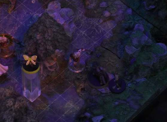 A battlemap of a sparse forested space focused on Thimble flying above Wulferic, who faces off against two zombies. One of the zombies is large and carrying an axe. The other is thin and spindly. Another zombie with an orange condition ring and the edge of Teor’s cape are visible at the edge of the image.