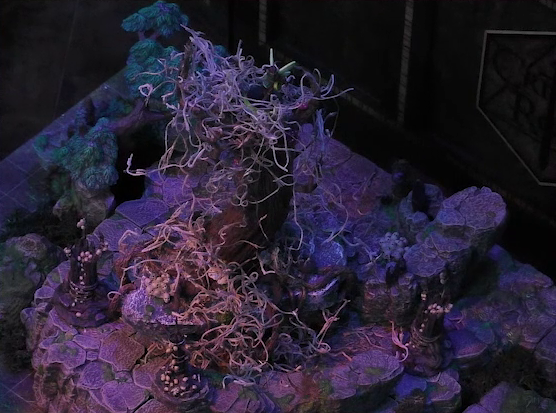 A battlemap of the Wraith Tree on a large rocky mound in a sparse forested space. Thimble stands on top of the tree. The Wraith Tree has thin, white, spaghetti-like tendrils hanging from its bare branches. Three small wooden structures covered in skulls surround the tree.