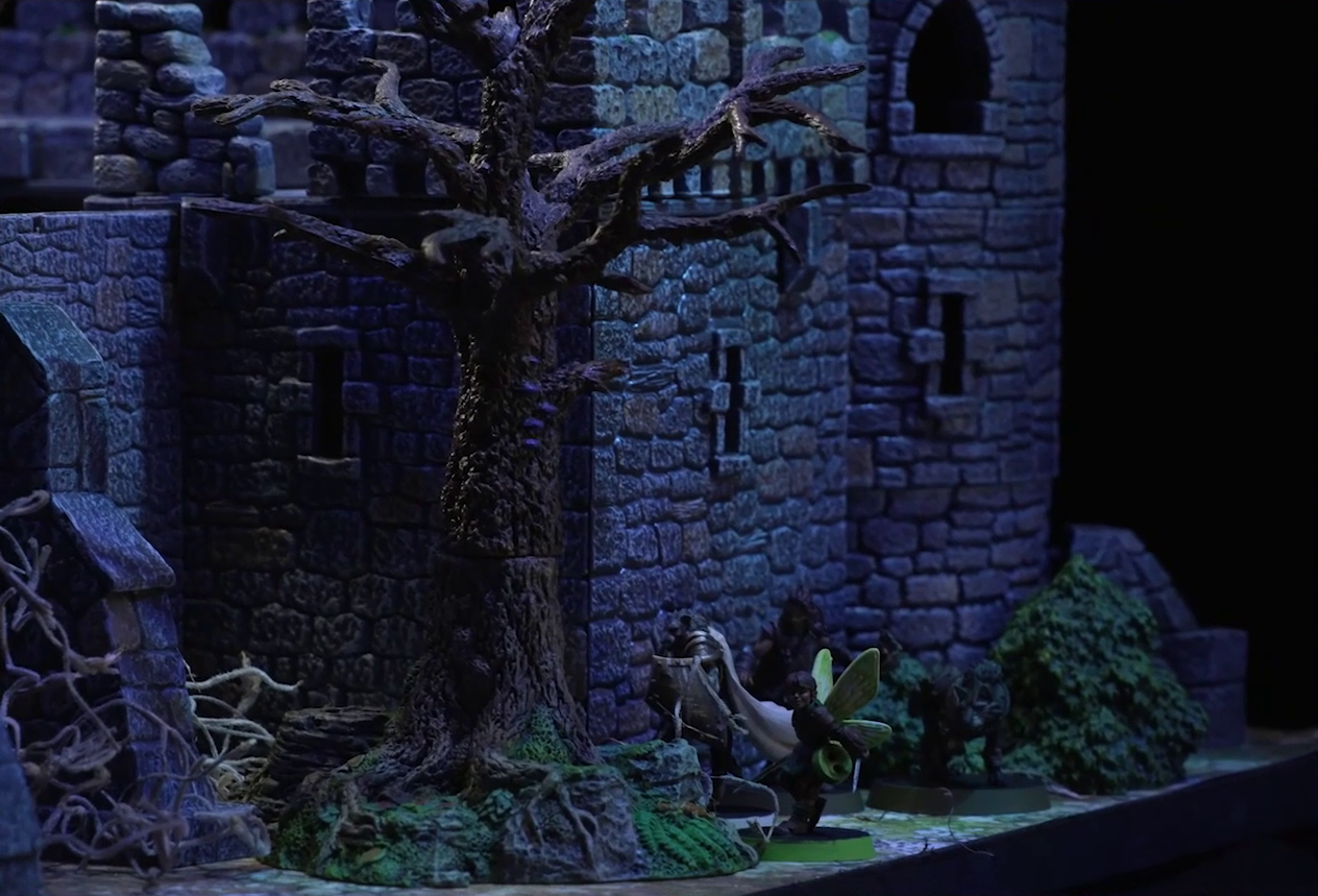 A battlemap of a stone castle focused on Thimble, Teor, Kattigan, and Wulferic standing by an exterior wall. A dead leafless tree rises nearby, and thin tangled roots emerge from the stonework. Empty windows are set in the wall above the party.