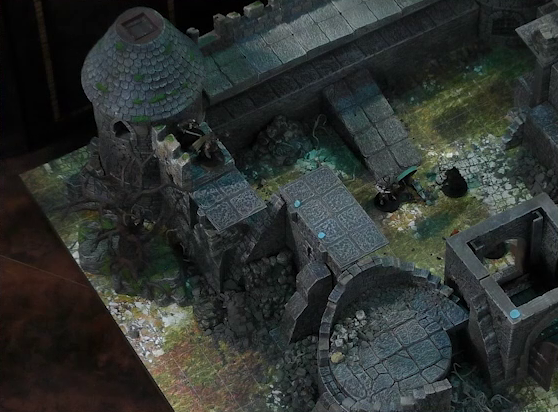 A battlemap of a ruined stone castle focused on Kattigan facing off against an armored guard on a courtyard wall. The tower on one end of that wall has collapsed. Inside the courtyard two individuals stand over a fallen Wicander. The wooden entry door into the courtyard is open.