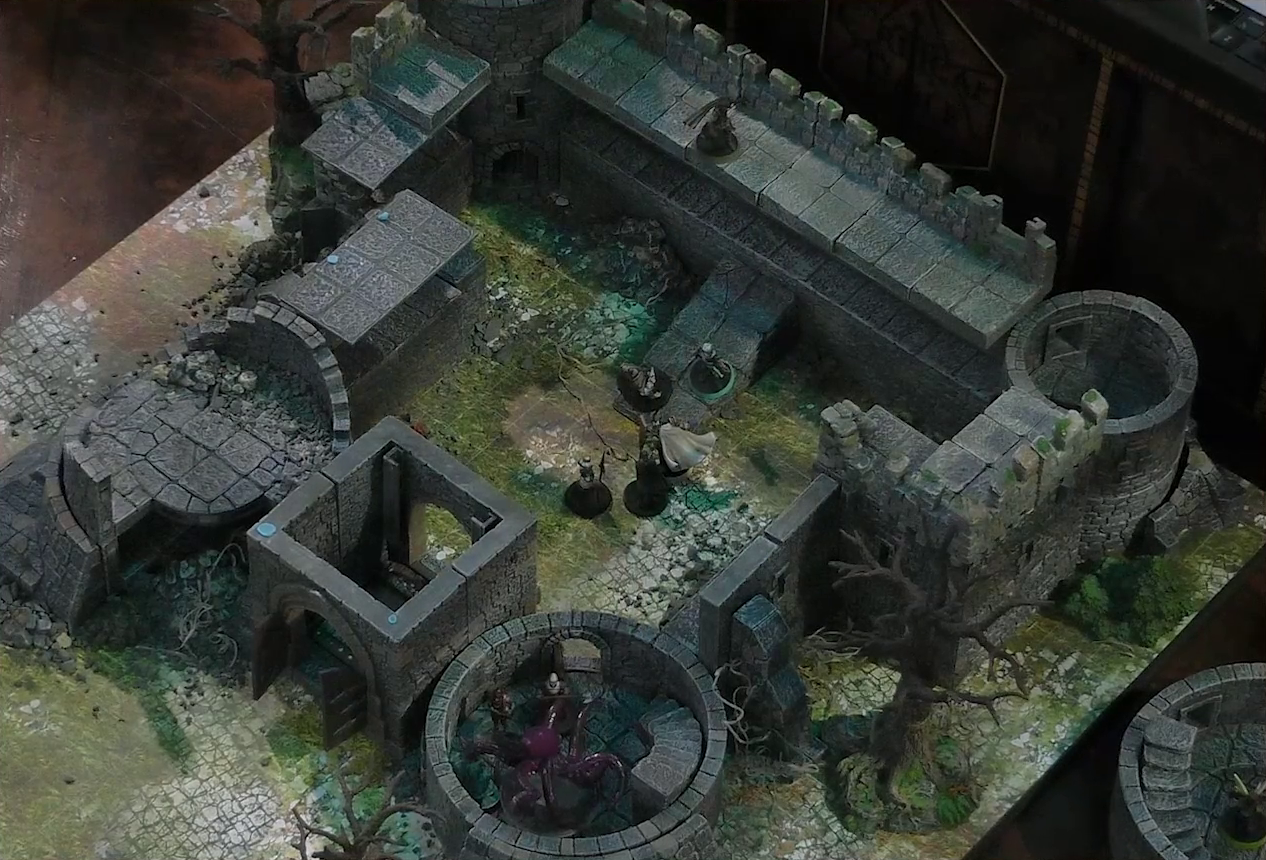 A battlemap of a ruined stone castle with sparse dead trees around its edges. The walled courtyard has a tower at each corner. A giant red squid is in the center of the bottom right tower along with Tyranny and some armored individuals. Wicander and Teor are in the courtyard facing off against three armored knights. Kattigan overlooks the courtyard from the rear wall. The bottom-left tower of the courtyard has collapsed.