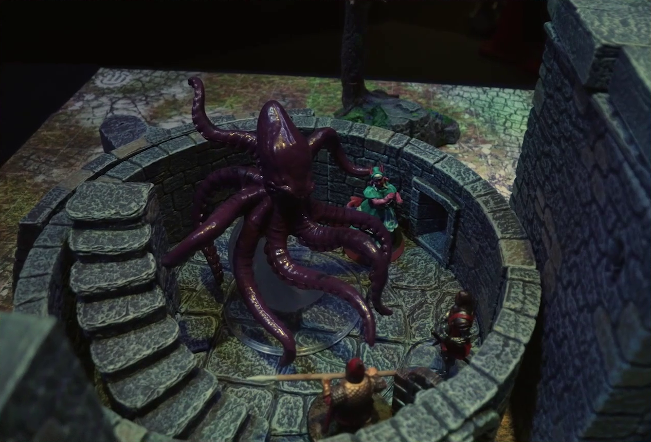 A battlemap of a ruined stone castle focused on a giant red squid in the interior of a small circular tower room. Tyranny stands to the side of the creature, while two armored soldiers face off against it, one wielding a spear and the other without a visible weapon. A staircase curves up along the edge of the room.