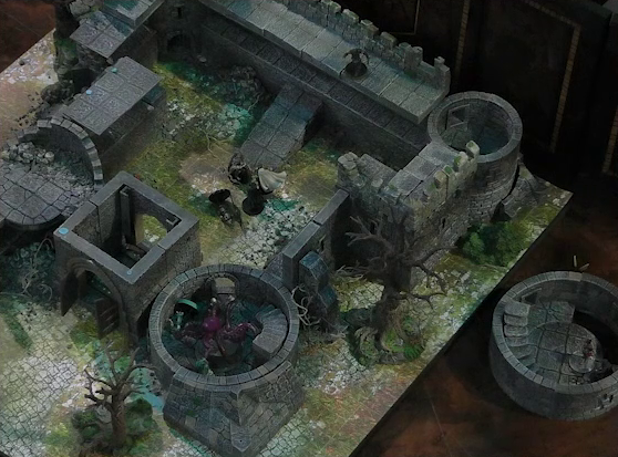 A battlemap of a ruined stone castle with sparse dead trees around its edges. The walled courtyard has a tower at each corner. A giant red squid is in the bottom-right tower along with Tyranny and a fallen Wicander. Teor faces off against a number of combatants in the courtyard, one fallen. Kattigan looks down over the courtyard from the rear wall. A knight stands in a tower room placed off to the side of the main battlemap. The bottom-left tower has collapsed.