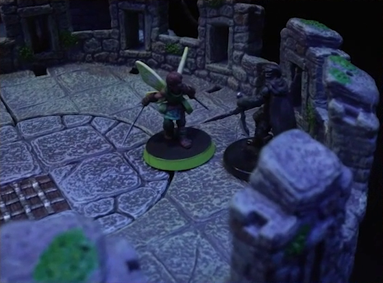 A battlemap of a stone parapet tower top, where Thimble faces off against Casimir. Thimble’s wings are outstretched, her needle swords spread out at her sides. Casimir, backed up against the edge, is dressed in black with a cape, and holds a dagger point down. A grate is in the center of the tower, and moss grows on the tops of the parapets.