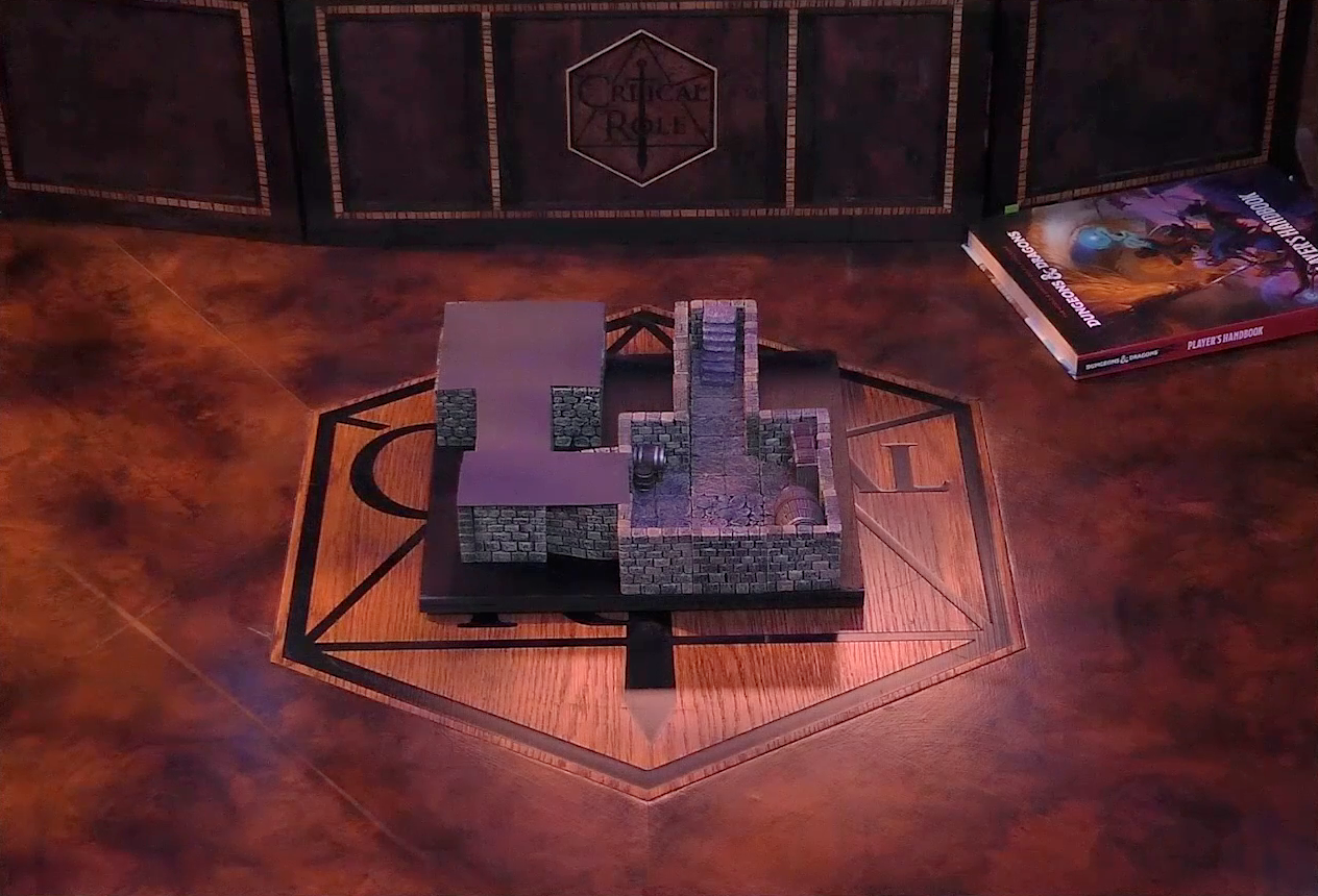A battlemap of a small stone dungeon. A short set of stairs go down into a hallway which leads to a chamber containing some barrels against the walls. Another hallway leads off and turns a corner into a second chamber. Both this final hallway and chamber are obscured with black construction paper. The battlemap sits on the table over the Critical Role logo. Behind it is the Critical Role DM screen and a copy of the D&D Player’s Handbook.
