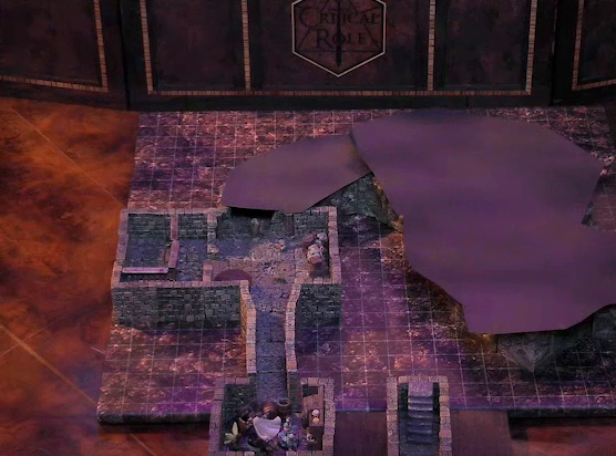 A battlemap of a stone dungeon. Wicander, Tyranny, Wulferic, Kattigan, Teor, and Thimble are in a small chamber with barrels against the walls. The chamber has entrances on opposite sides. One entrance leads into a hallway which opens up into a larger chamber with some wooden containers in the corner and a table opposite them. On one end of this chamber are metal bars resembling an empty jail cell. The chamber continues into another hallway and larger space which are both obscured by black construction paper. A short set of stairs go down into a hallway in another part of the dungeon.