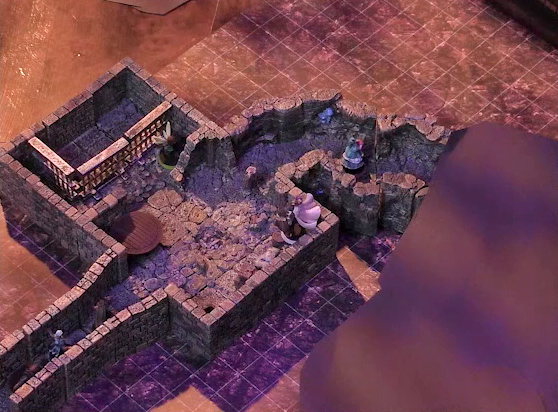 A battlemap of a stone dungeon focused on a chamber with hallways leading out of opposite ends of it. The chamber has some wooden containers in the corner and a table opposite them. On one end of the chamber are metal bars resembling an empty jail cell. Wulferic, Wicander, and Kattigan walk into the chamber via one hallway while Tyranny walks down the opposite hallway, which appears to be more naturally-formed. Teor stands in the corner near the wooden containers. Thimble stands near the jail cell.