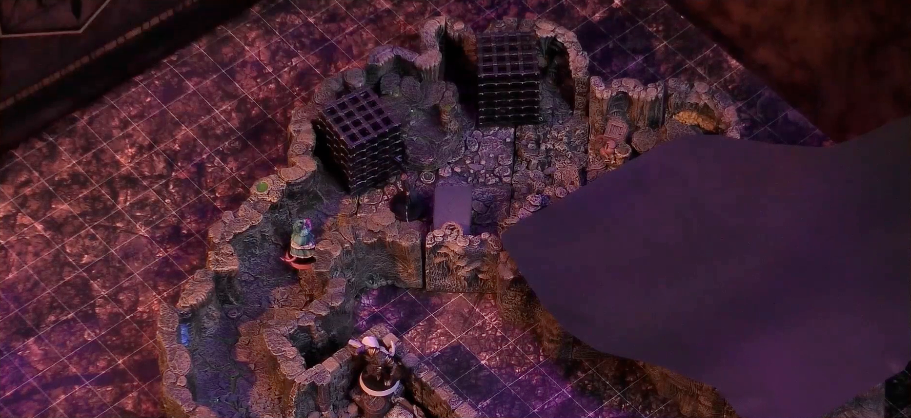 A battlemap of a stone dungeon focused on a naturally-formed chamber with two large black cube-shaped cages and a black stone slab. A figure wearing black stands between the slab and one cage. Tyranny enters the chamber from a rough-hewn stone hallway. Teor is further back along Tyranny’s hallway, standing in the corner of a more man-made stone chamber among some wooden containers and bags. Further in the room with the cages are some stone crates and jars.