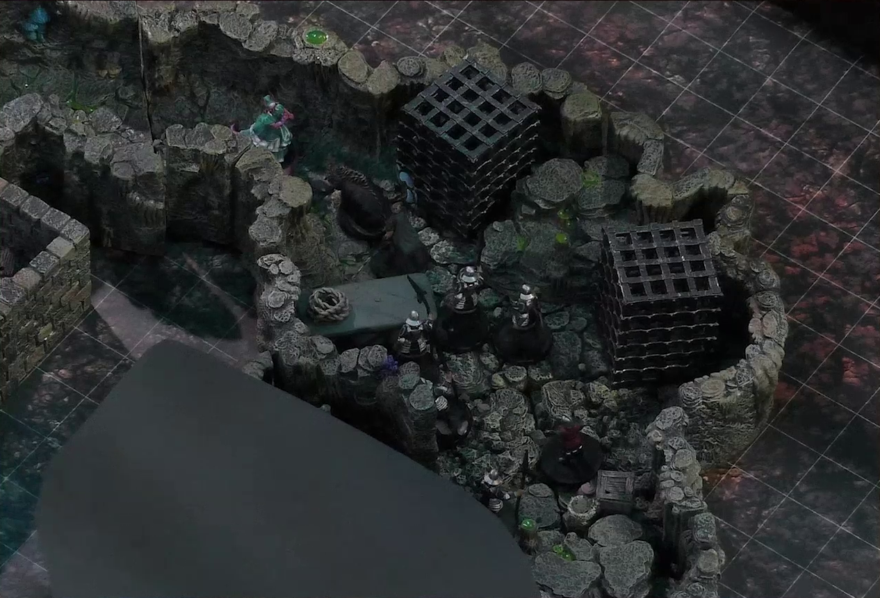 A battlemap of a naturally-formed small stone dungeon chamber. Tyranny faces off against a basilisk, a black-cloaked knight, and five armored guards. Two large black cube-shaped cages are against one wall, and a stone slab with a coil of rope on it is against the opposite wall. The area deeper into the dungeon is obscured by black construction paper. Small glowing green dots and turquoise mushrooms are scattered across the floor and walls.