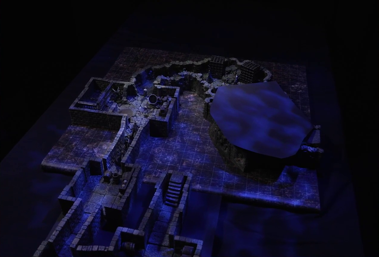 A battlemap of a stone dungeon. Stairs lead down into a series of man-made hallways and small chambers, eventually opening up into a larger naturally-formed space. The dungeon past this space is obscured with black construction paper. Wulferic, Wicander, and Kattigan stand single-file in a narrow hallway. Thimble is in the next room, by metal bars resembling an empty jail cell. Teor lies downed in the corner across from her. Deeper into the dungeon is a naturally-formed chamber, where Tyranny faces off against a basilisk, a black-cloaked knight, and some armored guards. Two large black cube-shaped cages are against one wall.