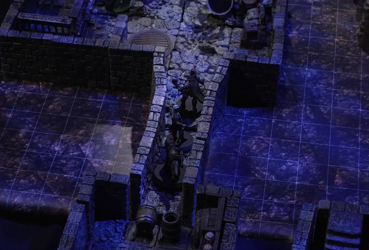 A battlemap of a stone dungeon focused on Wulferic, Wicander, and Kattigan lined up single-file in a narrow hallway. Behind them is a small chamber with barrels lining the walls and two barrels blocking the entry to the hall. In front of them is a larger chamber with some wooden containers against the wall and a round table opposite them. Teor lies downed near the containers, while Thimbel stands near metal bars resembling an empty jail cell against another wall.
