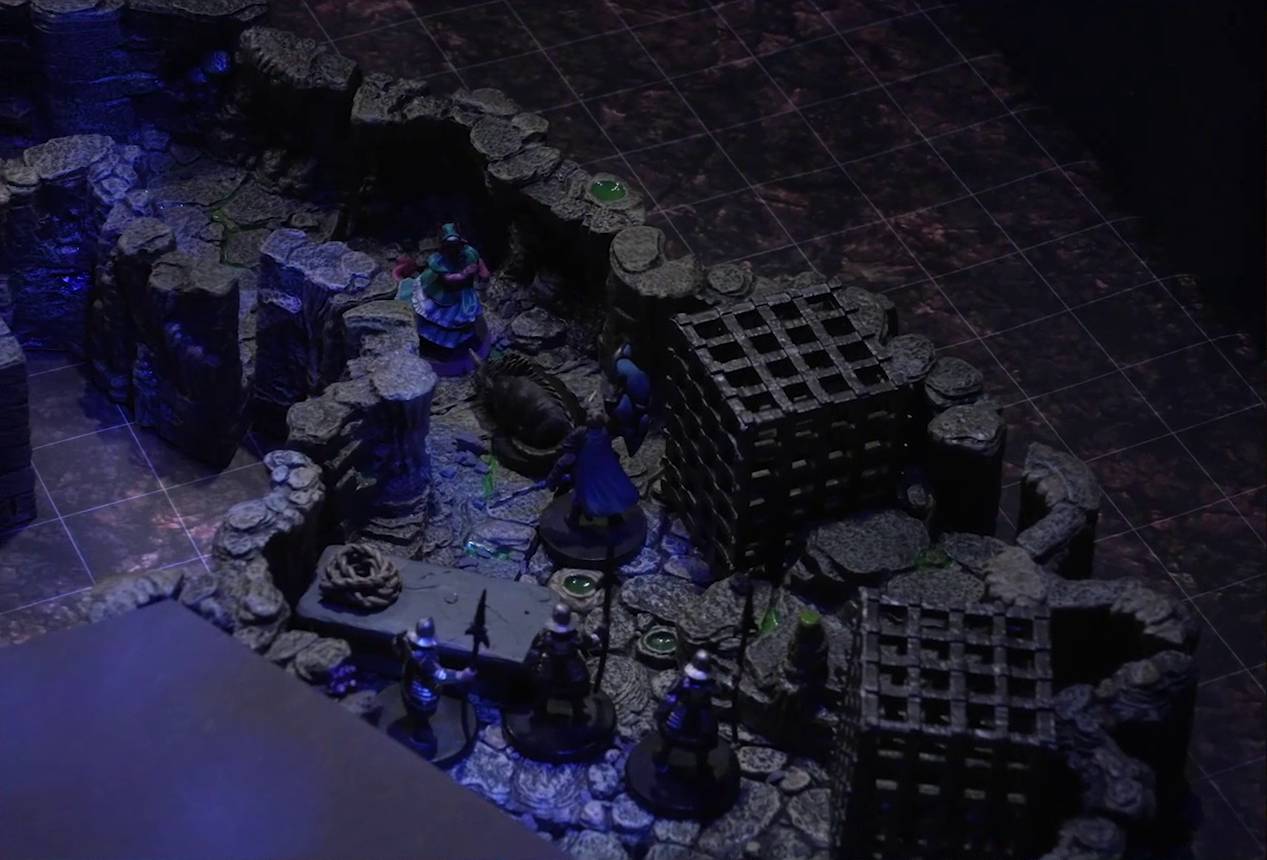 A battlemap of a naturally-formed small stone dungeon chamber. Tyranny faces off against a basilisk, a black-cloaked knight, and three armored guards with spears. Two large black cube-shaped cages are against one wall, and a stone slab with a coil of rope on it is against the opposite wall. The area deeper into the dungeon is obscured by black construction paper. Small glowing green dots and turquoise mushrooms are scattered across the floor and walls.