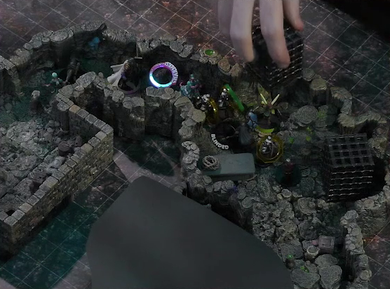 A battlemap of a naturally-formed small stone dungeon chamber. Brennan’s hand reaches down to lift a large black cube-shaped cage off of Thimble. Another cage is next to it and a stone slab with a coil of rope on it is against the opposite wall. Wulferic, Wicander, Teor, Kattigan, and Tyranny face off against a basilisk, a black-cloaked knight, and some armored guards. The guards have yellow status rings on them, Kattigan has a “CHARMED” status ring on him, and the basilisk has a “BLINDED” status on it. The area deeper into the dungeon is obscured by black construction paper.