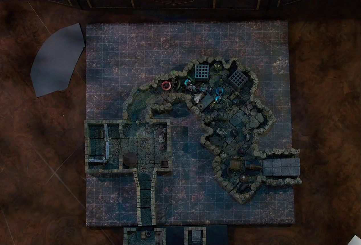 A top-down view of a battlemap of a stone dungeon, revealing a newly uncovered section where a horse-drawn-cart is set to exit upwards via a ramp, guarded by a few soldiers. Some statues stand near the cart. Kattigan, Wulferic, Wicander, Tyranny, Teor, and Thimble face off against a black-cloaked knight and some armored guards in a crowded naturally-formed chamber. Two large black cube-shaped cages are against one wall, and a stone slab with a coil of rope on it is against the opposite wall. Kattigan has a “CHARMED” status ring on him. Further back in the dungeon is a man-made room with metal bars resembling an empty jail cell and some wooden containers in the corner.