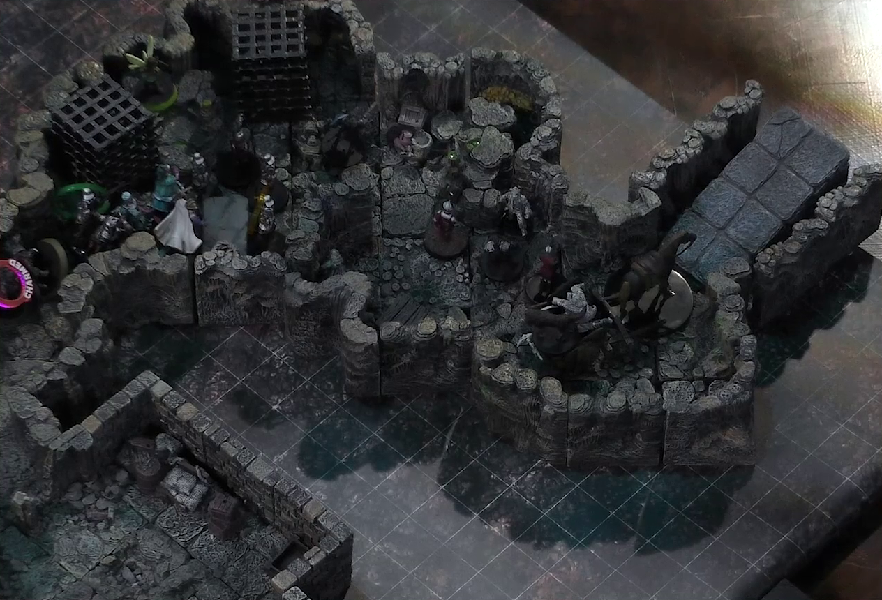 A battlemap of a naturally-formed small stone dungeon chamber. A cart piled high with statues, both of people and of a dog, is pulled by a horse towards a ramp exiting upwards. Another statue stands by the wall near the cart. A number of armored guards stand by the cart. Further back, Kattigan, Wicander, Tyranny, Wulferic, and Teor face off against a black-cloaked knight and some more armored guards. Kattigan has a “CHARMED” status ring on him. Two large black cube-shaped cages are against one wall, and a stone slab with a coil of rope on it is against the opposite wall.