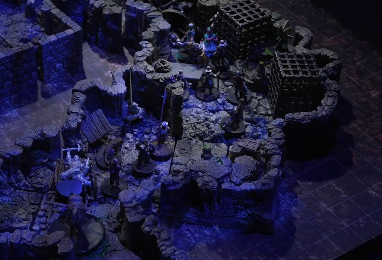 A battlemap of a naturally-formed small stone dungeon chamber, bathed in blue light. A cart bearing a statue of a person wielding a mining pick is pulled by a horse towards a ramp exiting upwards. A statue of a dog stands near the cart. A number of armored guards stand by the cart. Further back, Wulferic, Wicander, Tyranny, and Thimble face off against a black-cloaked knight and some more armored guards. Two large black cube-shaped cages are against one wall, and a stone slab with a coil of rope on it is against the opposite wall.