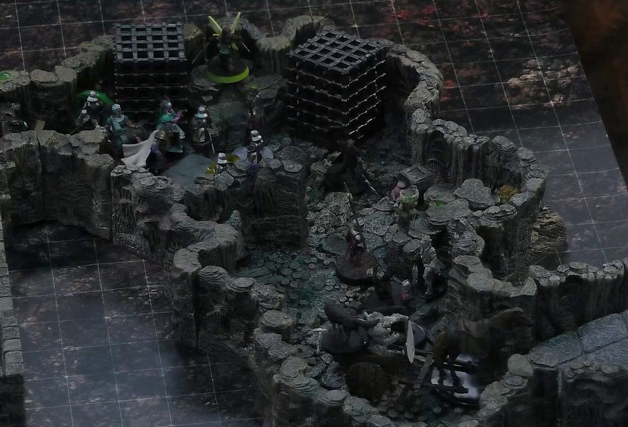 A battlemap of a naturally-formed small stone dungeon chamber. A cart piled high with statues, both of people and of a dog, is pulled by a horse towards a ramp exiting upwards. Another statue stands by the wall near the cart. A number of armored guards stand by the cart. Further back, Kattigan, Wicander, Tyranny, Wulferic, Teor, and Thimble face off against a black-cloaked knight and some more armored guards. Two large black cube-shaped cages are against one wall, and a stone slab with a coil of rope on it is against the opposite wall.