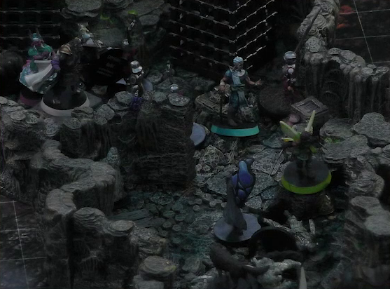 A battlemap of a naturally-formed small stone dungeon chamber. Wicander stands in the middle of the action, facing off against a black-cloaked knight and some armored guards alongside Tyranny, Teor, and Thimble. Thimble stands over a fallen statue. Two large black cube-shaped cages are against one wall, and a stone slab on which Teor stands is against the opposite wall.