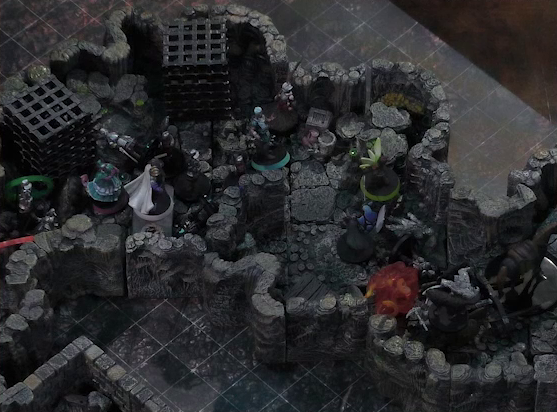 A battlemap of a naturally-formed small stone dungeon chamber. A flaming cart piled high with statues, both of people and of a dog, is pulled by a horse towards a ramp exiting upwards. Another statue lays on the ground by the wall near the cart, at Thimble’s feet. A number of armored guards stand by the cart. Further back, Kattigan, Wicander, Tyranny, Wulferic, Teor, and Thimble face off against a black-cloaked knight and some more armored guards. Two large black cube-shaped cages are against one wall, and a stone slab with a coil of rope on it is against the opposite wall.