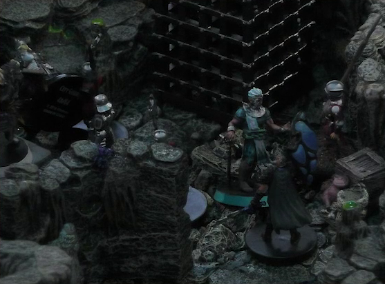 A battlemap of a naturally-formed small stone dungeon chamber. Wicander stands in the middle of the action, facing off against a black-cloaked knight and some armored guards. Two large black cube-shaped cages are against one wall, and a stone slab on which Teor stands is against the opposite wall.