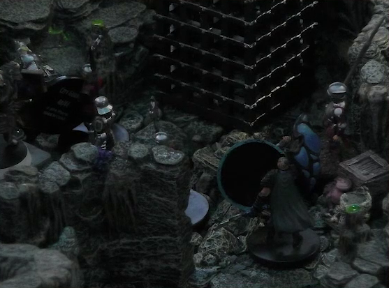 A battlemap of a naturally-formed small stone dungeon chamber. Wicander lies on the ground near a black-cloaked knight and some armored guards. Two large black cube-shaped cages are against one wall, and a stone slab on which Teor stands is against the opposite wall.