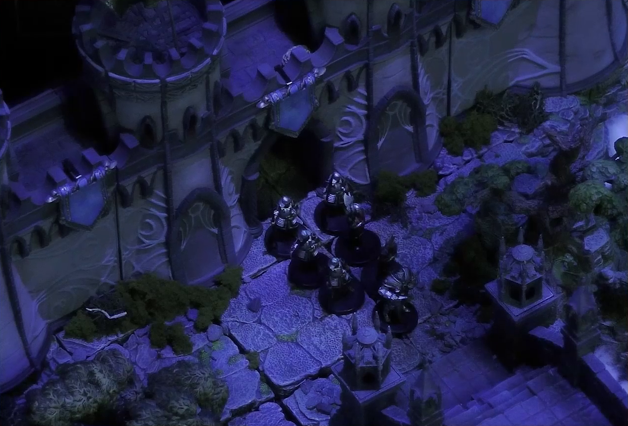 A battlemap of the end of an ornate stone bridge leading to a stone wall with parapets and towers. Six armored soldiers led by Frons Tachonis stand before a gate in the wall.