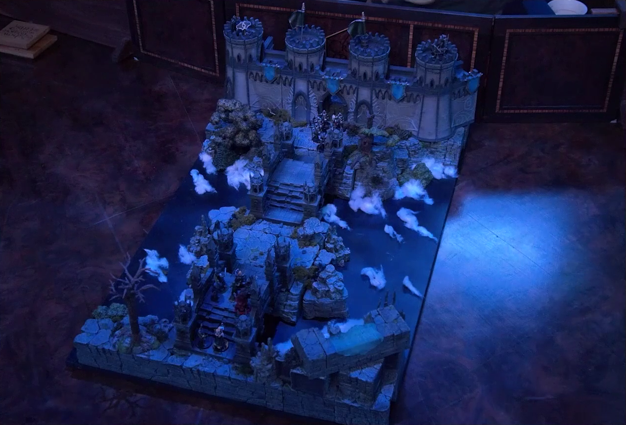 A battlemap of an ornate stone bridge over water leading to a stone wall with parapets and towers. Julien, Mozor, Vaelus, Occtis, Aranessa, and Thaisha stand at one end, while six armored soldiers led by Frons Tachonis stand before a gate in the wall at the other end. Two ballistas are atop the towers. White foam caps waves in the water below.