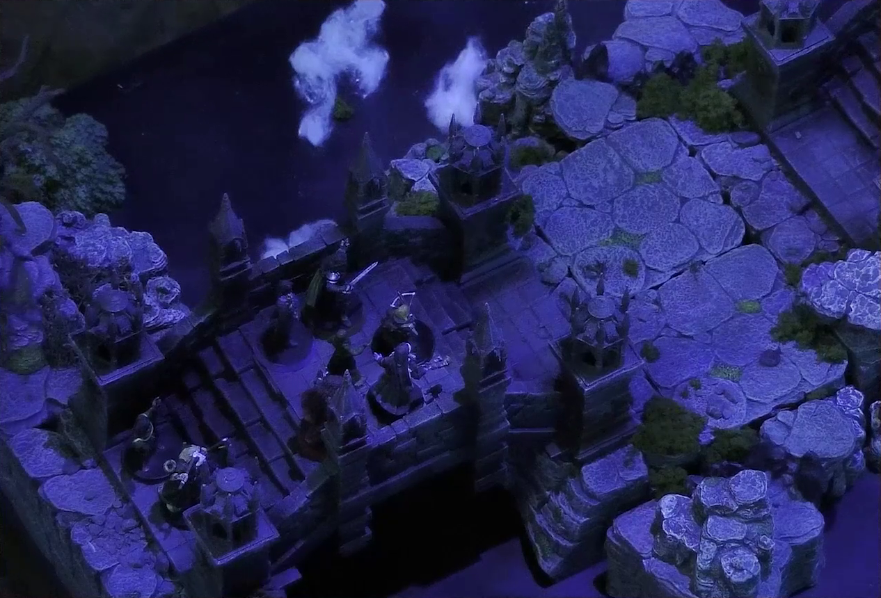 A battlemap of an ornate stone bridge over water. Julien, Mozor, Vaelus, Occtis, Aranessa, and Thaisha stand at one end. Behind them are two druids. White foam caps waves in the water below.