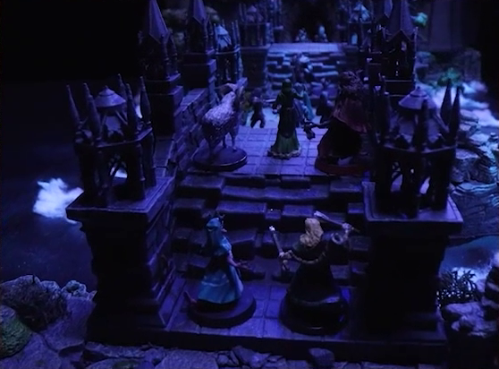 A battlemap of an ornate stone bridge over water leading to a stone wall. Cruelty and an axe-wielding druid are in the foreground at the edge of the bridge, while Hala, Aranessa, and Thaisha stand further along the bridge. Julien, Enmity, Vaelus, and Occtis face off against the heavily-armored Frons Tachonis further along still, with six armored soldiers following behind Frons.