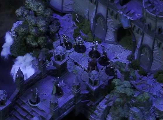 A battlemap of an ornate stone bridge over water leading to a stone wall with parapets and towers. Six armored soldiers form a line blocking Thaisha from reaching a gate through the wall at the end of the bridge. Just behind Thaisha are Julien and Agony. White foam caps waves in the water below.