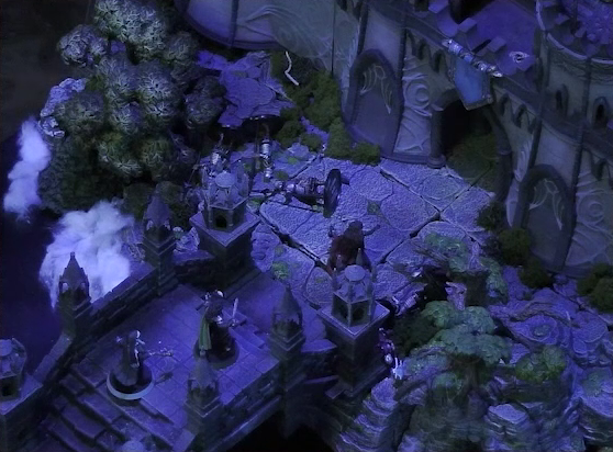 A battlemap of an ornate stone bridge over water leading to a stone wall with parapets and towers. Six armored soldiers lay on the ground between Thaisha and a gate through the wall at the end of the bridge. Just behind Thaisha are Julien and Agony. White foam caps waves in the water below.
