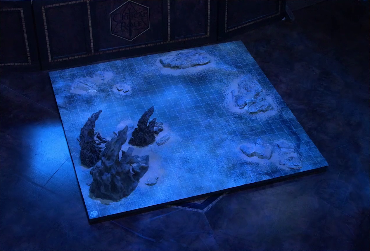 A battlemap of a flat, sandy expanse under the cover of night. Sparse jagged rocky formations jut from the otherwise empty landscape. The battlemap sits on the Critical Role table in front of the DM screen.