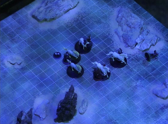 A battlemap of a flat, sandy expanse under the cover of night. Julien lays on the ground some distance away from the party’s white horses. Vaelus stands by the horse closest to him. Occtis, Pin, Thaisha, and Gaya Seremai stand in a circle with the four other horses. Some jagged rocky formations jut from the otherwise empty landscape.