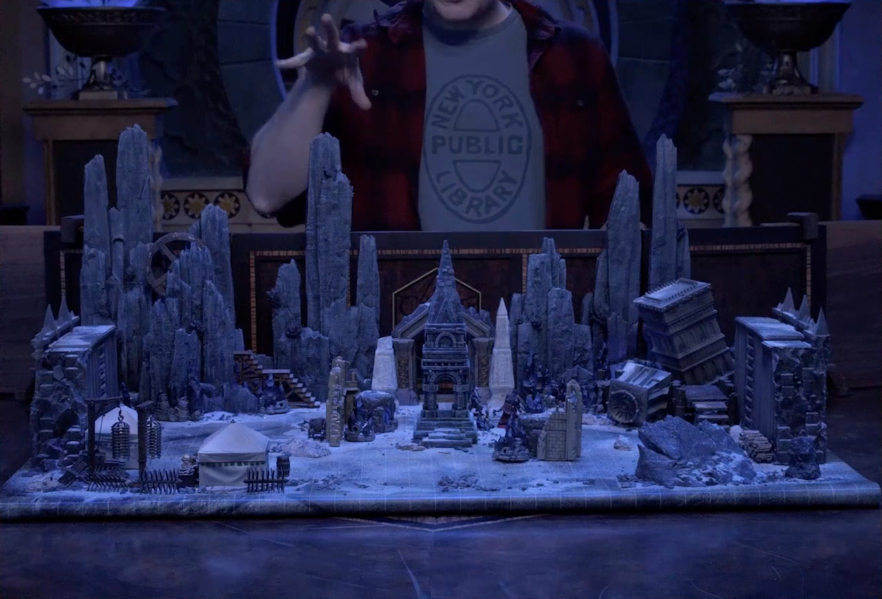 A battlemap of stony desert ruins under the cover of night. A short tower stands in the middle of the battlemap among fallen walls and collapsed towers. A camp with two square tents, palisades, and hanging iron cages is off to the side. A gate is at the rear of the battlemap, with tall jutting rocks on either side. Some wooden stairs lead up these rocks towards a large wooden wheel. Brennan is in the background behind the DM screen, motioning over the battlemap with his right hand. He wears a New York Public Library T-shirt under a flannel button-up.